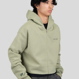 A model wearing a sage green zip-up hoodie with 'est. 1976' printed on the left chest and a 'tiff' logo on the right sleeve, paired with dark gray trousers, against a solid light gray background.
