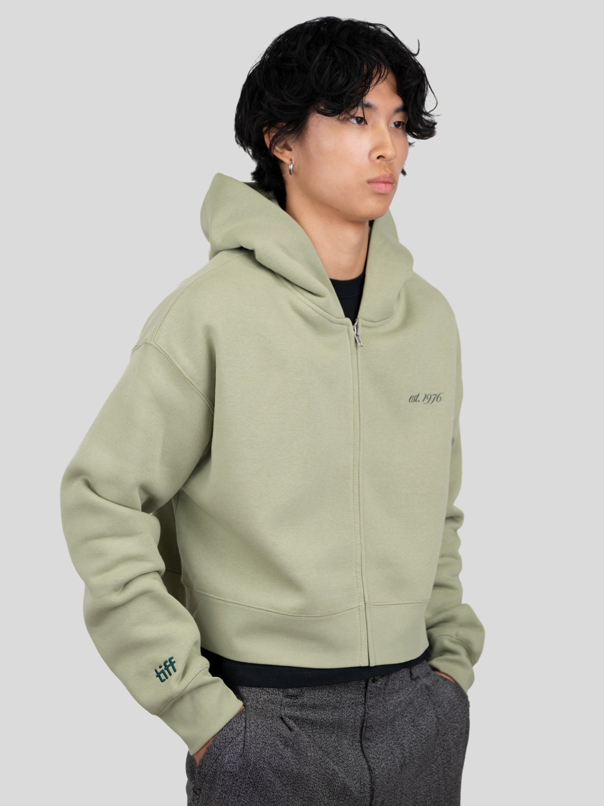 A model wearing a sage green zip-up hoodie with 'est. 1976' printed on the left chest and a 'tiff' logo on the right sleeve, paired with dark gray trousers, against a solid light gray background.
