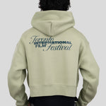 A person shown from the back wearing a light sage green cropped hoodie with a dark blue graphic print reading 'Toronto International Film Festival' on the back, paired with a black shirt and gray pants, against a solid light gray background.
