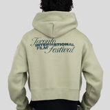 A person shown from the back wearing a light sage green cropped hoodie with a dark blue graphic print reading 'Toronto International Film Festival' on the back, paired with a black shirt and gray pants, against a solid light gray background.