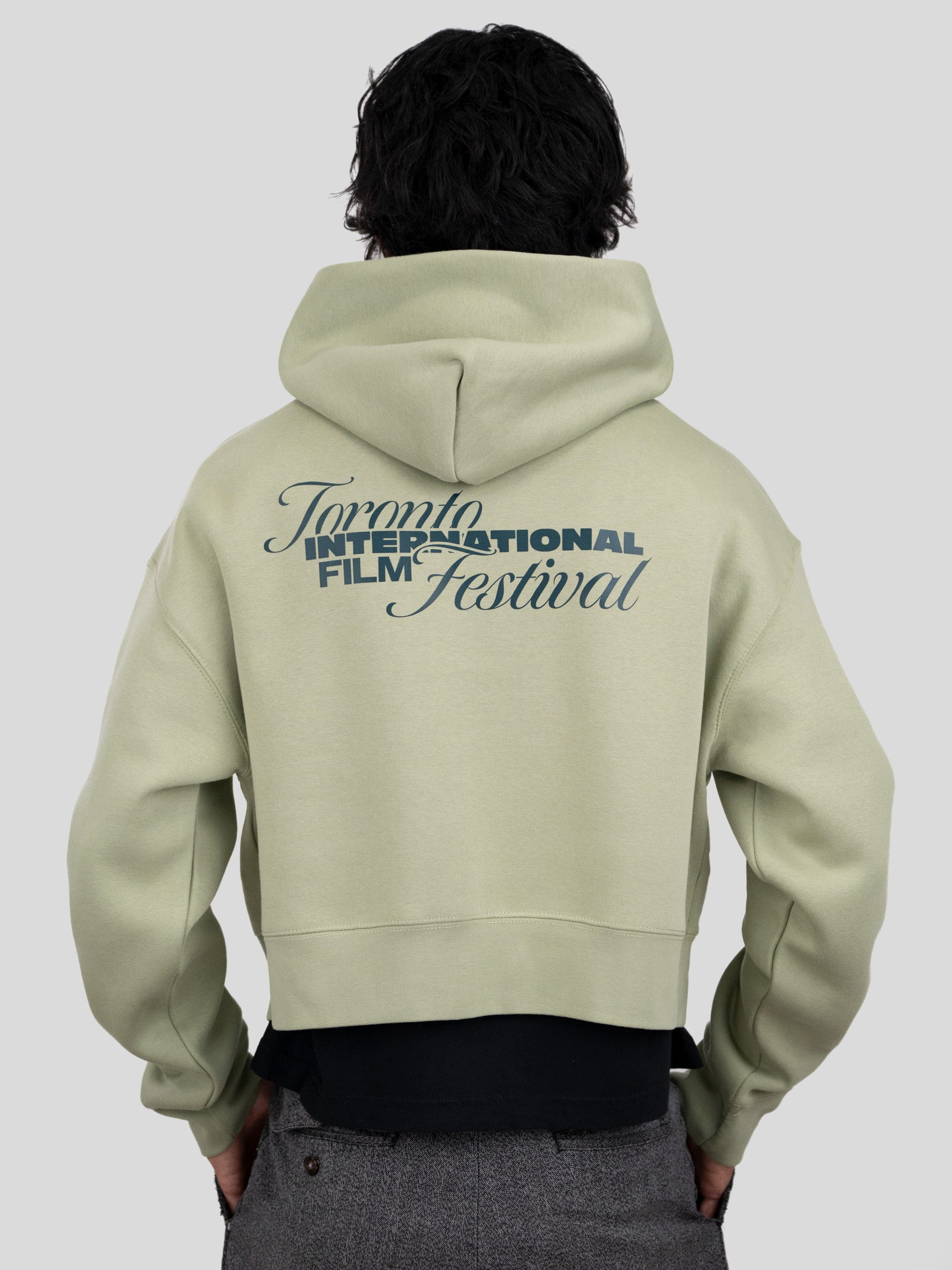 A person shown from the back wearing a light sage green cropped hoodie with a dark blue graphic print reading 'Toronto International Film Festival' on the back, paired with a black shirt and gray pants, against a solid light gray background.