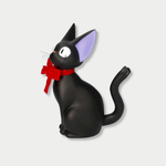 Side view of a black cat figurine representing Jiji from Kiki's Delivery Service sits upright with large eyes, purple inner ears, and a prominent red bow tied around its neck.


