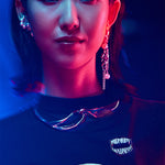 A person with slicked-back dark hair and pearl jewelry wears a black Midnight Madness t-shirt with a matching enamel pin, illuminated by high-contrast blue and magenta lighting.
