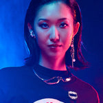 A person with slicked-back dark hair and pearl jewelry wears a black Midnight Madness t-shirt with a matching enamel pin, illuminated by high-contrast blue and magenta lighting.