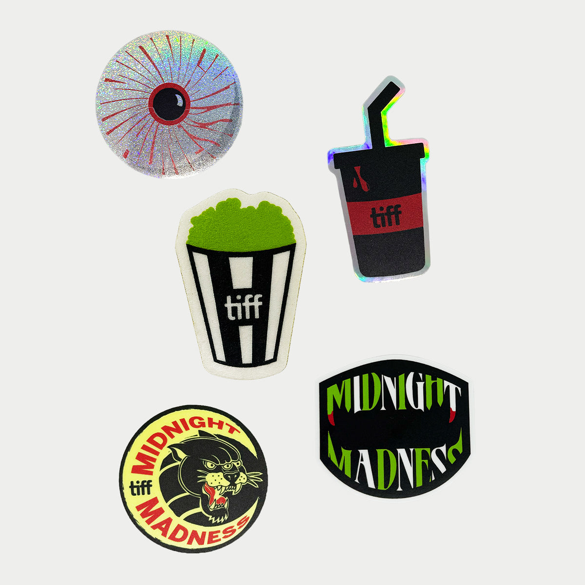 Group of five stickers with one glitter bloodshot eyeball, halographic black and red soda cup with straw, textured green popcorn with black and white striped bucket, green and white midnight madness logo with fangs, and round sticker with moon and panther graphic. 