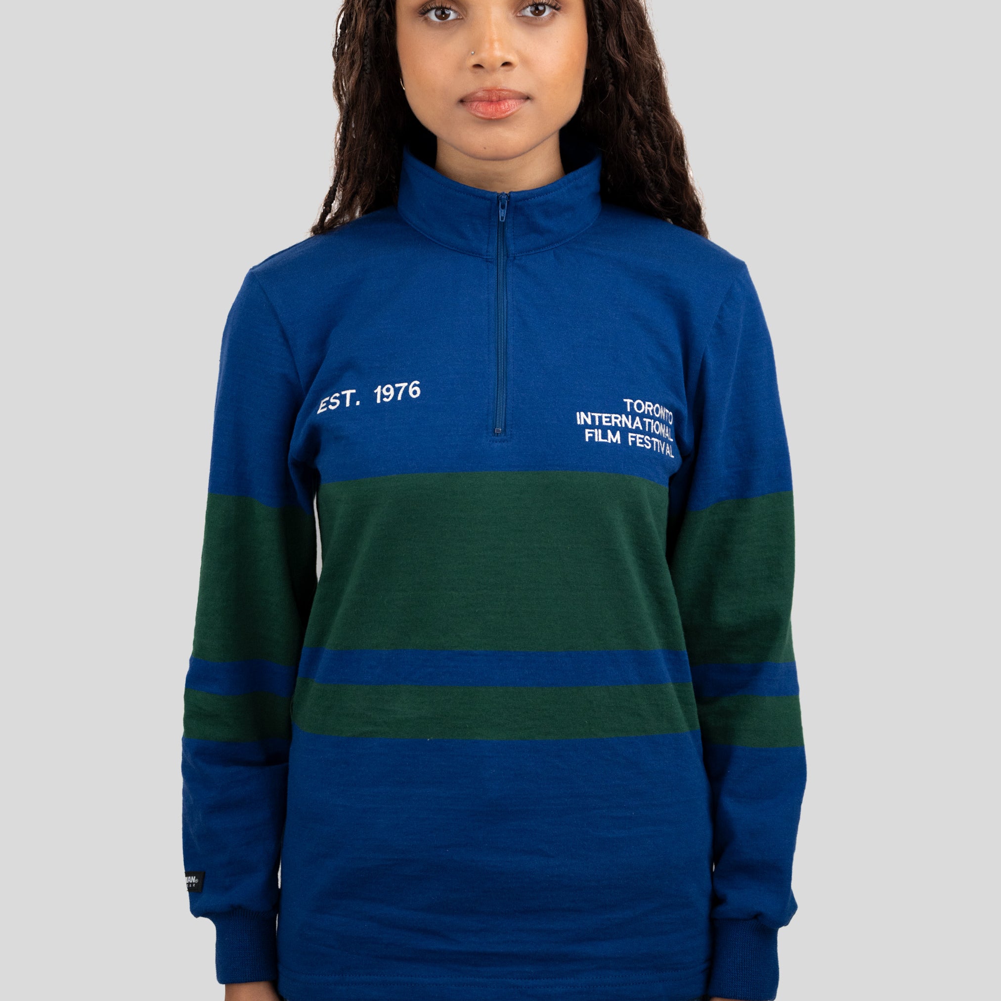 A model wears a blue and dark green quarter-zip pullover featuring "EST. 1976" and "TORONTO INTERNATIONAL FILM FESTIVAL" printed on the chest.