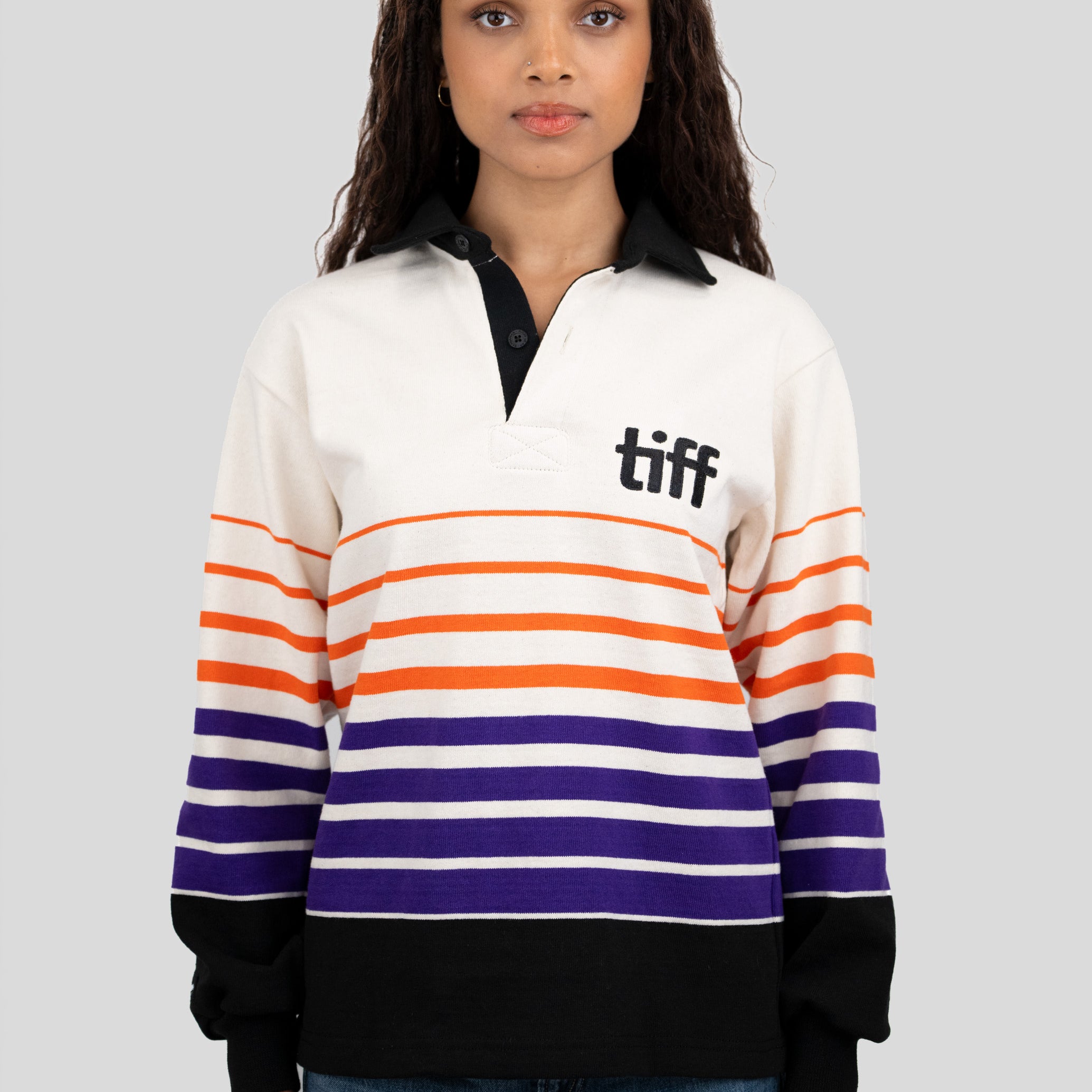 A model wears a long-sleeved, collared rugby shirt featuring orange, purple, and black stripes with the "TIFF" logo printed on the left chest.