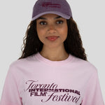 A woman wearing a light pink crewneck t-shirt and a dusty purple baseball cap, both featuring the 'Toronto International Film Festival' logo in dark purple. The photo is shot against a solid light gray background.