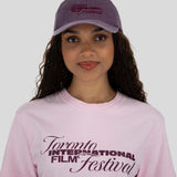 A woman wearing a light pink crewneck t-shirt and a dusty purple baseball cap, both featuring the 'Toronto International Film Festival' logo in dark purple. The photo is shot against a solid light gray background.