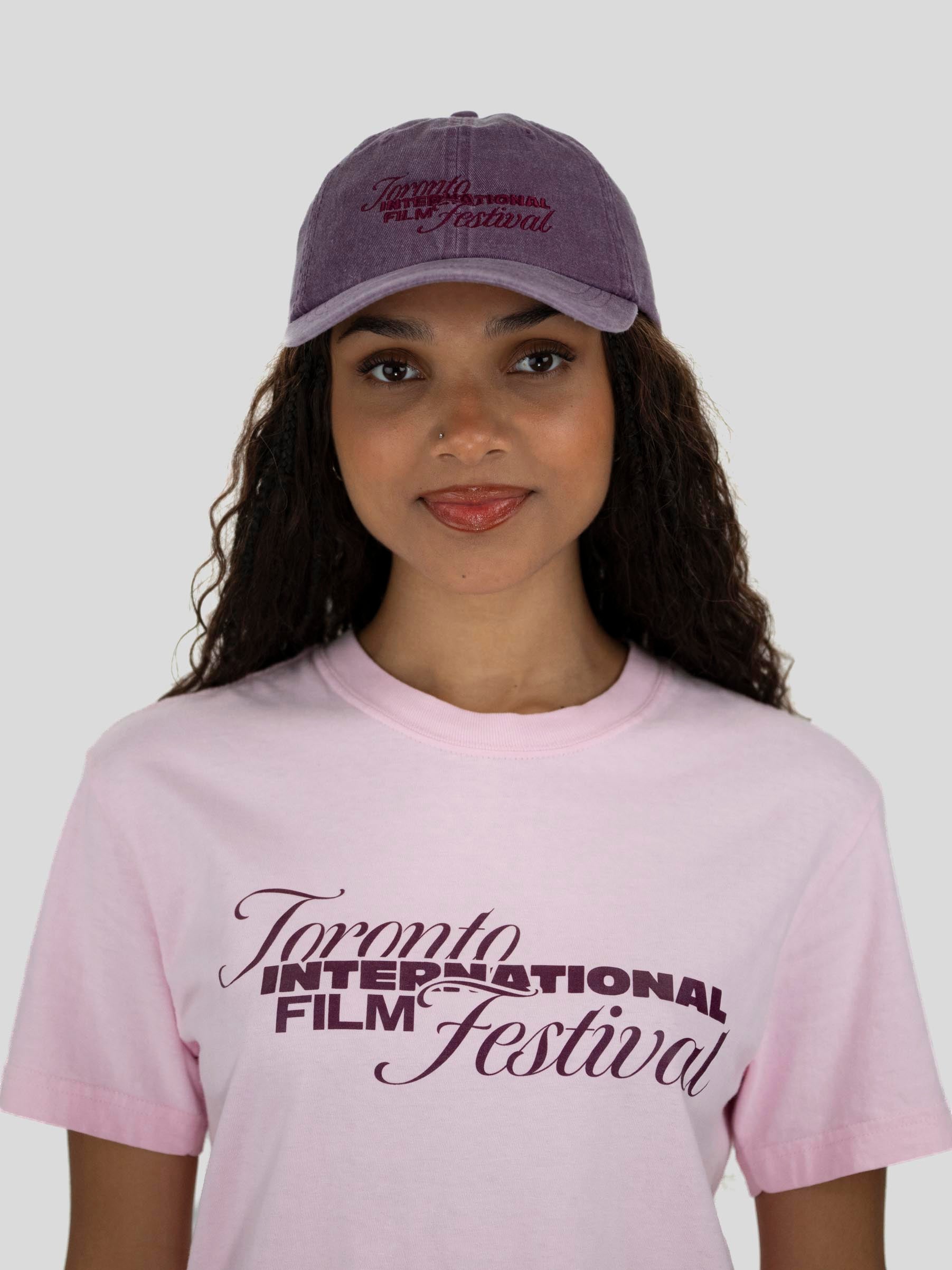 A woman wearing a light pink crewneck t-shirt and a dusty purple baseball cap, both featuring the 'Toronto International Film Festival' logo in dark purple. The photo is shot against a solid light gray background.