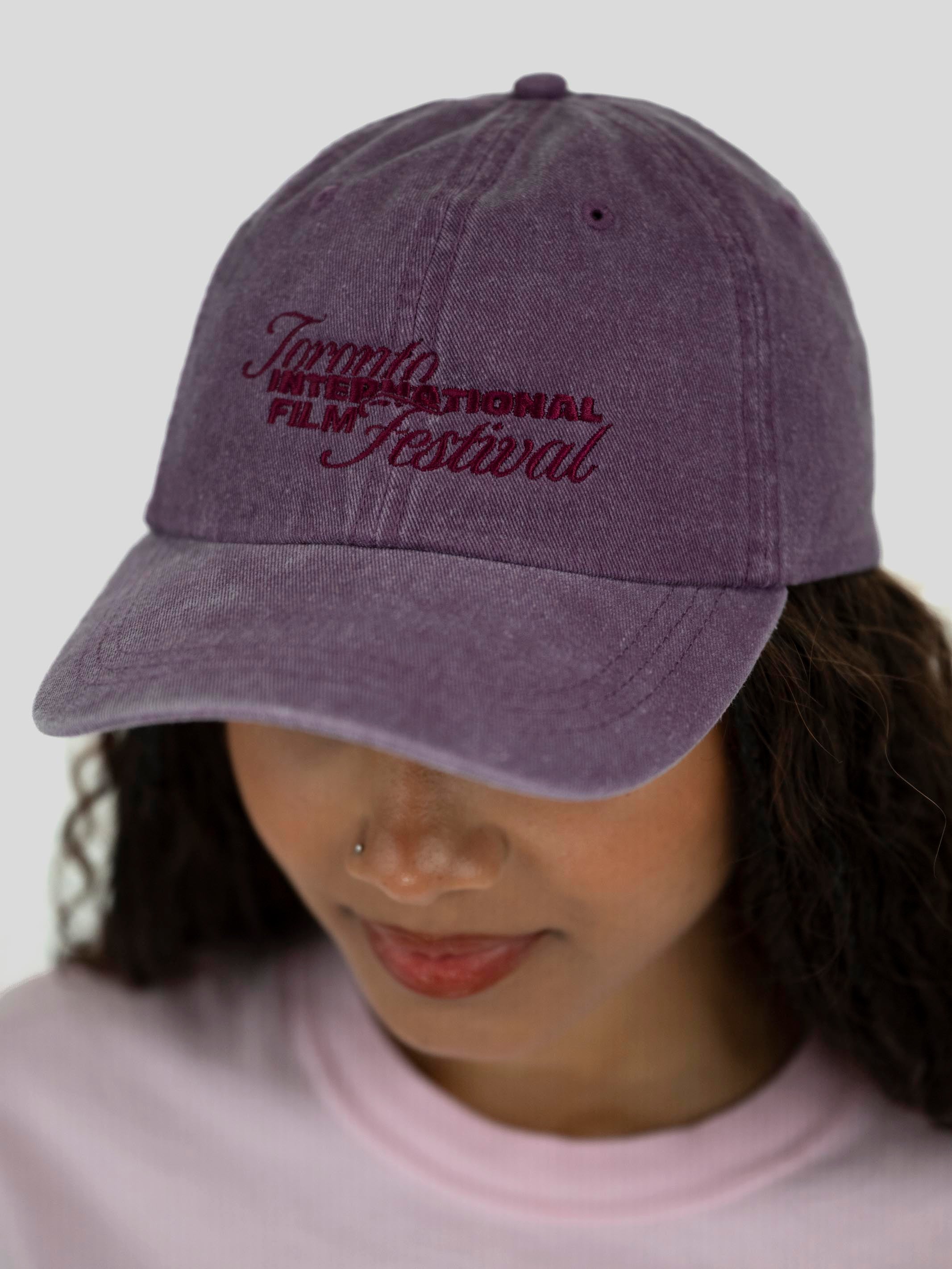 A person wearing a washed purple baseball cap with embroidered text reading 'Toronto International Film Festival' in dark red, shown against a light gray background.