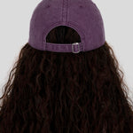A person viewed from the back wearing a purple washed baseball cap with a dark brown wavy wig, set against a solid light gray background.
