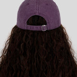 A person viewed from the back wearing a purple washed baseball cap with a dark brown wavy wig, set against a solid light gray background.