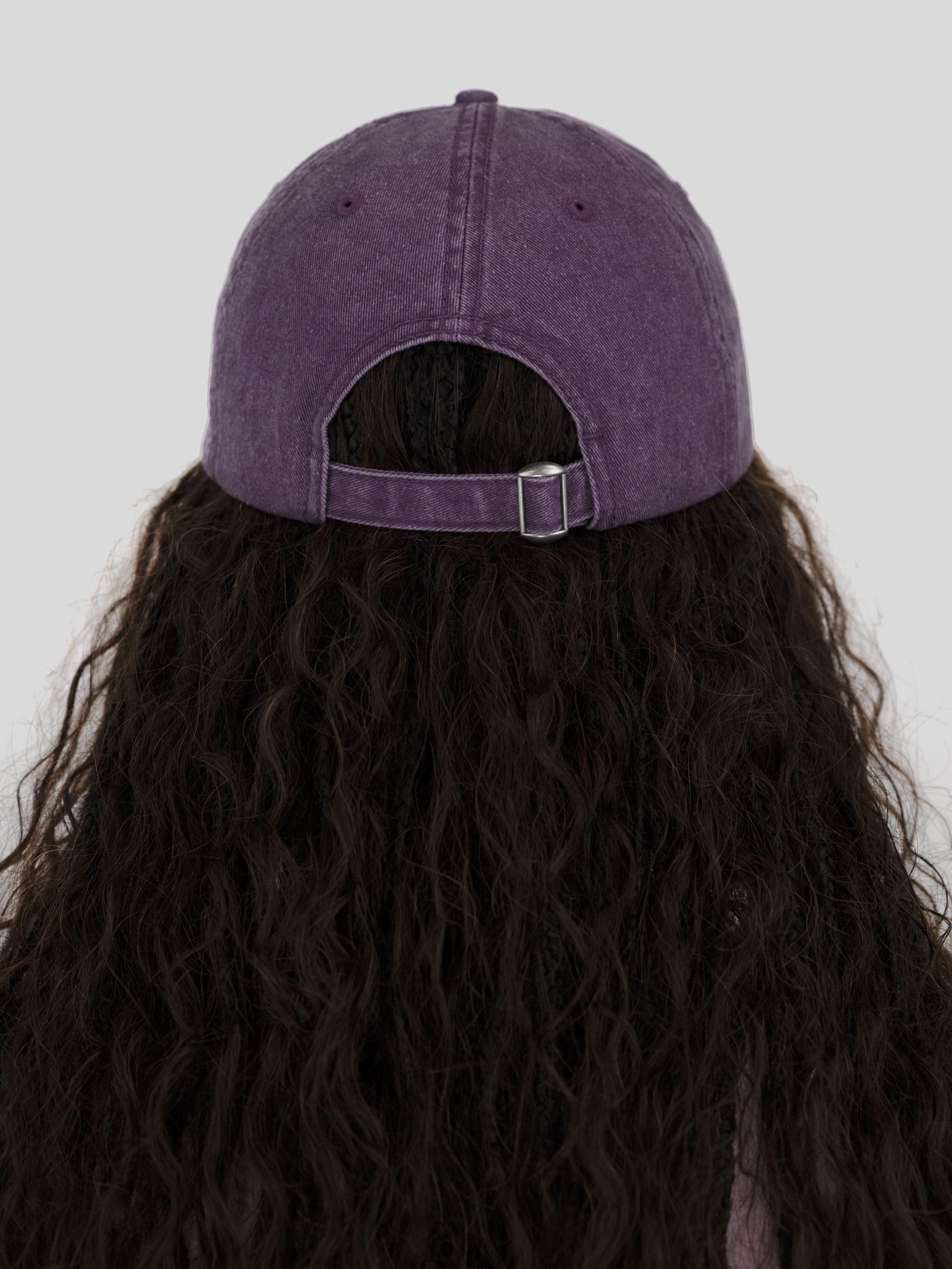 A person viewed from the back wearing a purple washed baseball cap with a dark brown wavy wig, set against a solid light gray background.