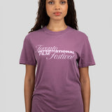 A woman wearing a purple short-sleeve crewneck t-shirt with 'Toronto International Film Festival' printed in white across the chest, paired with blue jeans, set against a solid light gray background.