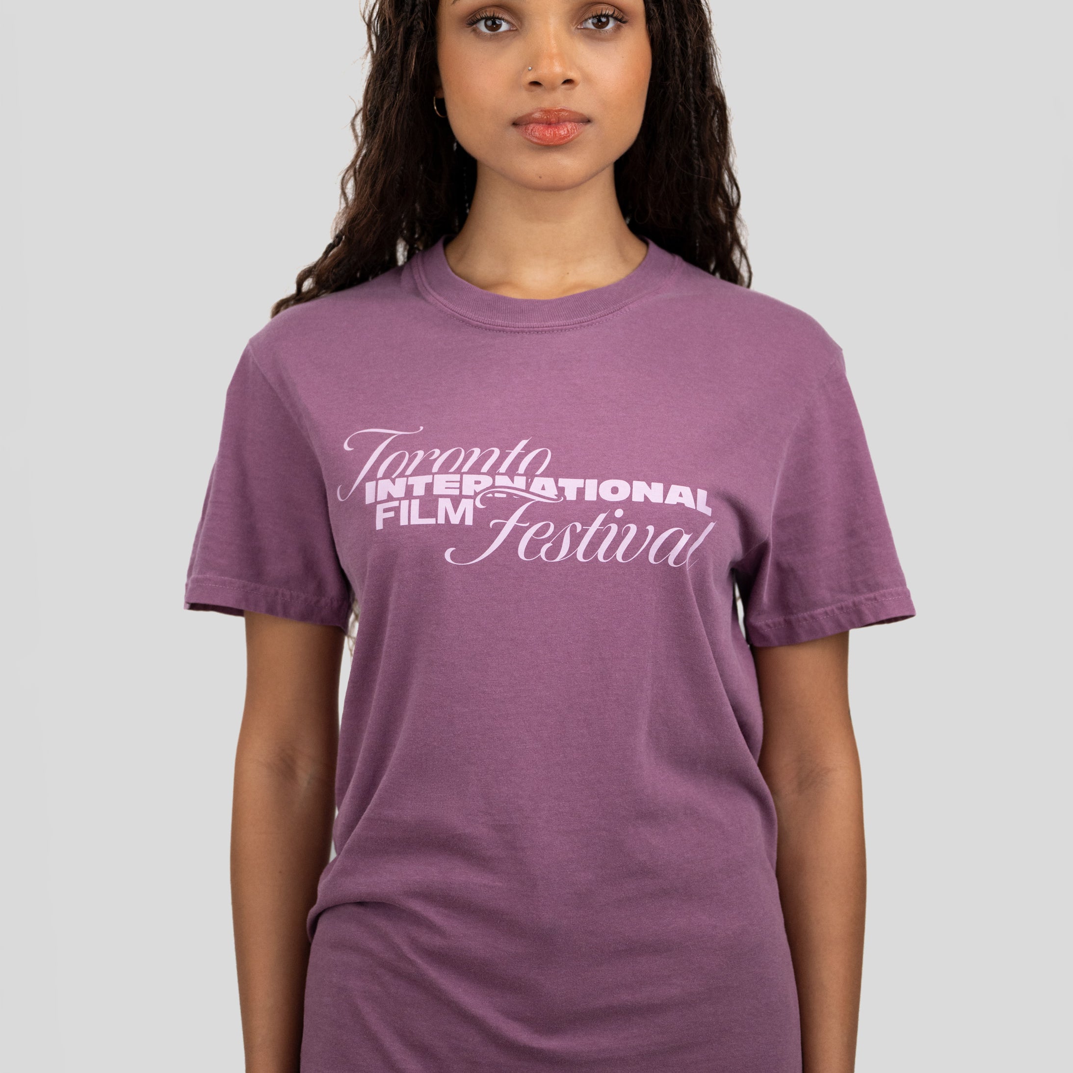 A woman wearing a purple short-sleeve crewneck t-shirt with 'Toronto International Film Festival' printed in white across the chest, paired with blue jeans, set against a solid light gray background.