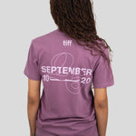 A woman viewed from behind wearing a mauve-purple T-shirt with a white graphic print on the back reading 'tiff SEPTEMBER 2026' and 'SEPTEMBER 10' with a line graphic. She is also wearing blue jeans against a solid light gray background.