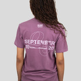 A woman viewed from behind wearing a mauve-purple T-shirt with a white graphic print on the back reading 'tiff SEPTEMBER 2026' and 'SEPTEMBER 10' with a line graphic. She is also wearing blue jeans against a solid light gray background.