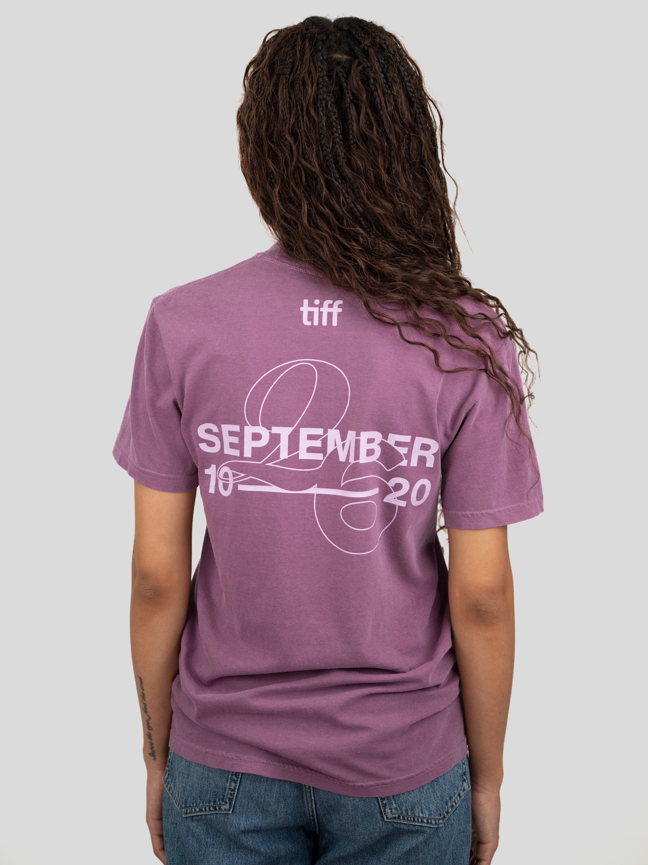 A woman viewed from behind wearing a mauve-purple T-shirt with a white graphic print on the back reading 'tiff SEPTEMBER 2026' and 'SEPTEMBER 10' with a line graphic. She is also wearing blue jeans against a solid light gray background.
