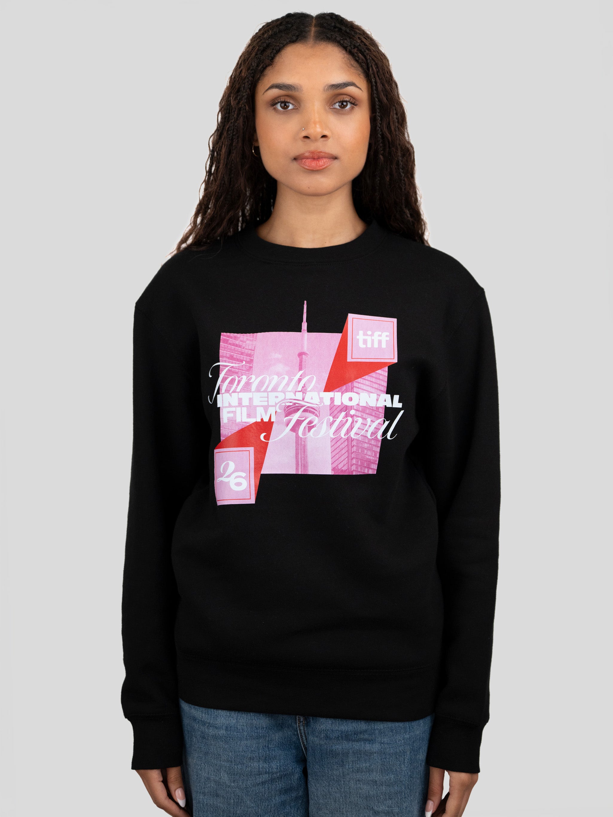 A woman modeling a black crewneck sweatshirt featuring a pink and red graphic print of the Toronto International Film Festival (TIFF) logo and building on the front, paired with blue jeans against a solid gray background.