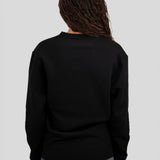 A person viewed from the back wearing a black long-sleeve crewneck sweater and blue jeans against a light gray studio background.