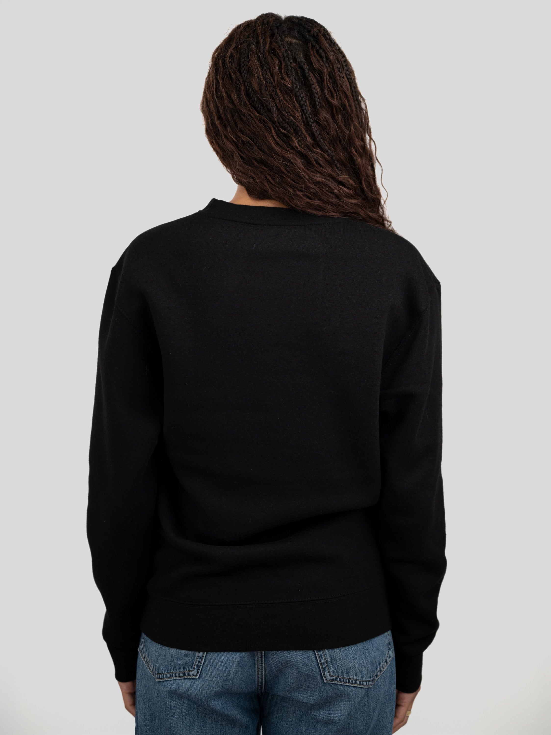 A person viewed from the back wearing a black long-sleeve crewneck sweater and blue jeans against a light gray studio background.