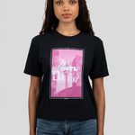 A woman wearing a black graphic t-shirt with a pink and white print, paired with blue jeans, against a solid gray background.