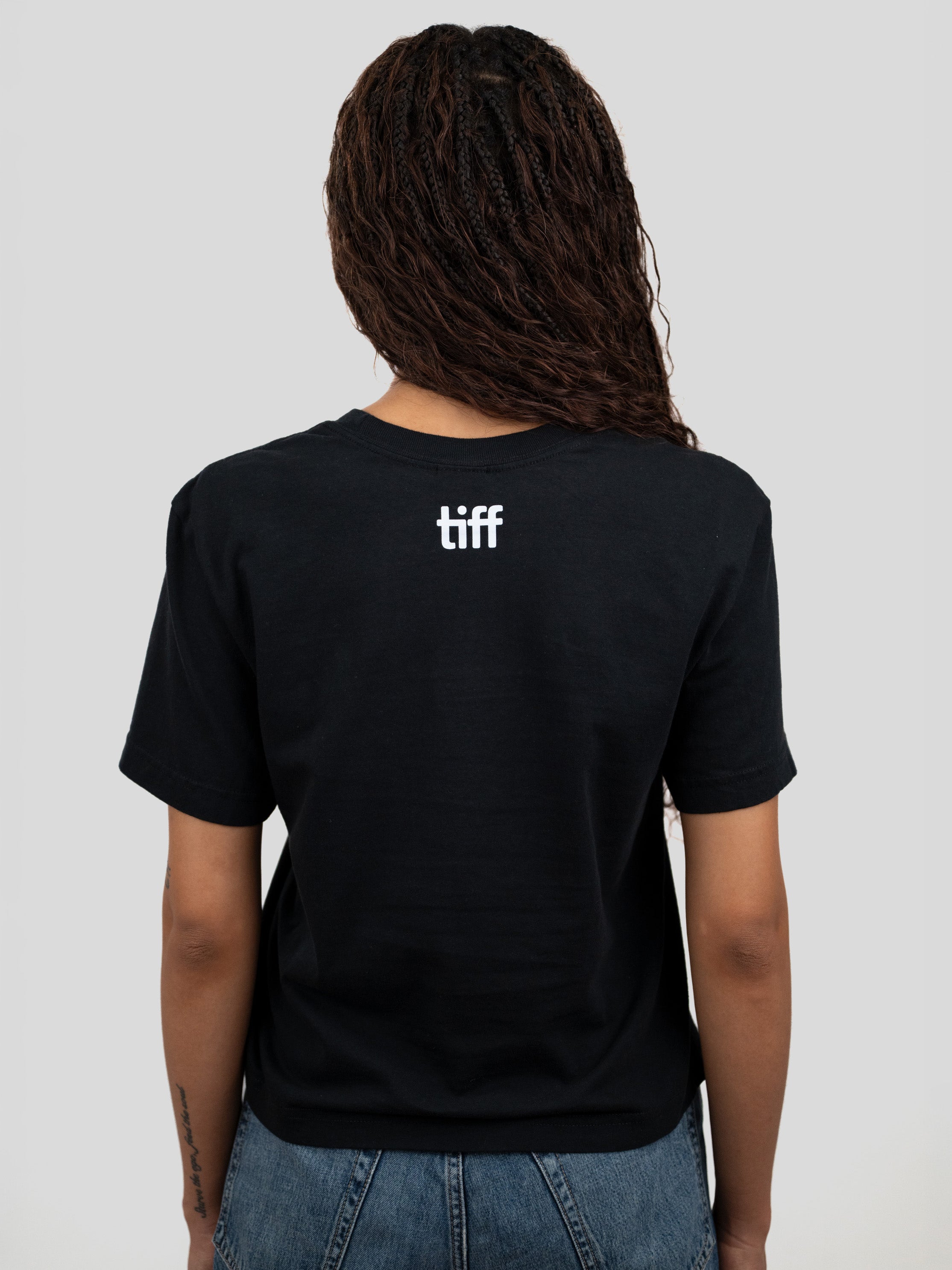 A person shown from the back wearing a black short-sleeve T-shirt with a small white 'tiff' logo centered on the upper back, paired with blue jeans against a light gray studio background.