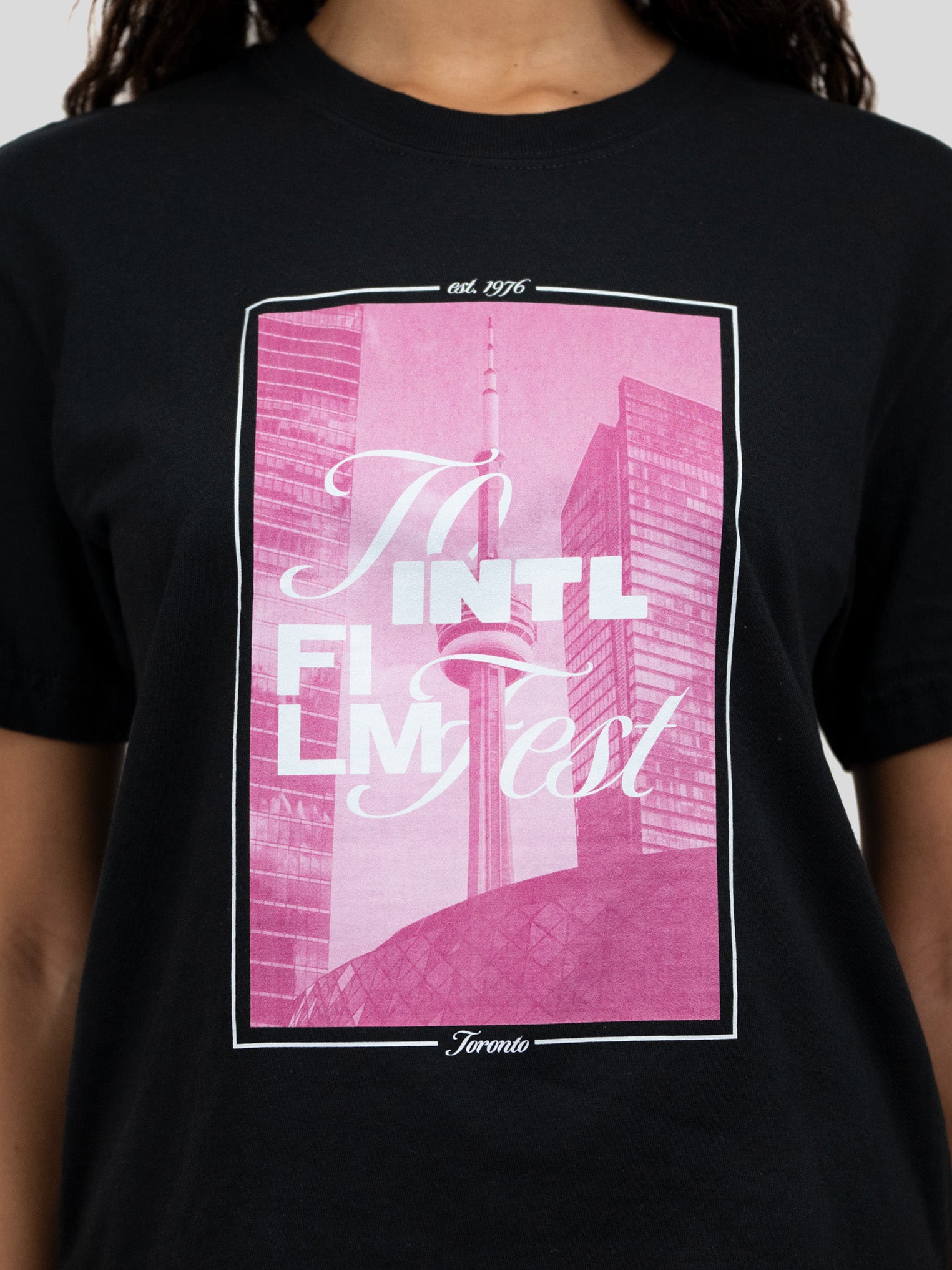 A black short-sleeve crewneck t-shirt featuring a large pink graphic print on the front with the text 'To Intl Film Fest' and a stylized Toronto skyline, worn by a model.