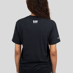 A woman viewed from behind wearing a black short-sleeve T-shirt with a white 'tiff' logo printed on the upper back, against a solid light gray background.