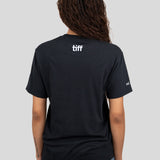 A woman viewed from behind wearing a black short-sleeve T-shirt with a white 'tiff' logo printed on the upper back, against a solid light gray background.