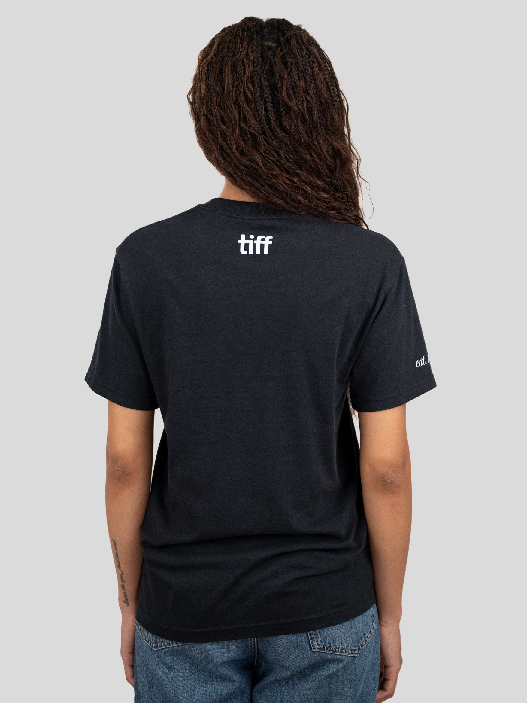 A woman viewed from behind wearing a black short-sleeve T-shirt with a white 'tiff' logo printed on the upper back, against a solid light gray background.