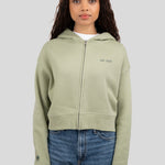 A woman modeling a light sage green cropped zip-up hoodie with 'est. 1976' embroidered on the left chest, paired with blue jeans, against a solid gray background.
