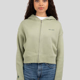 A woman modeling a light sage green cropped zip-up hoodie with 'est. 1976' embroidered on the left chest, paired with blue jeans, against a solid gray background.