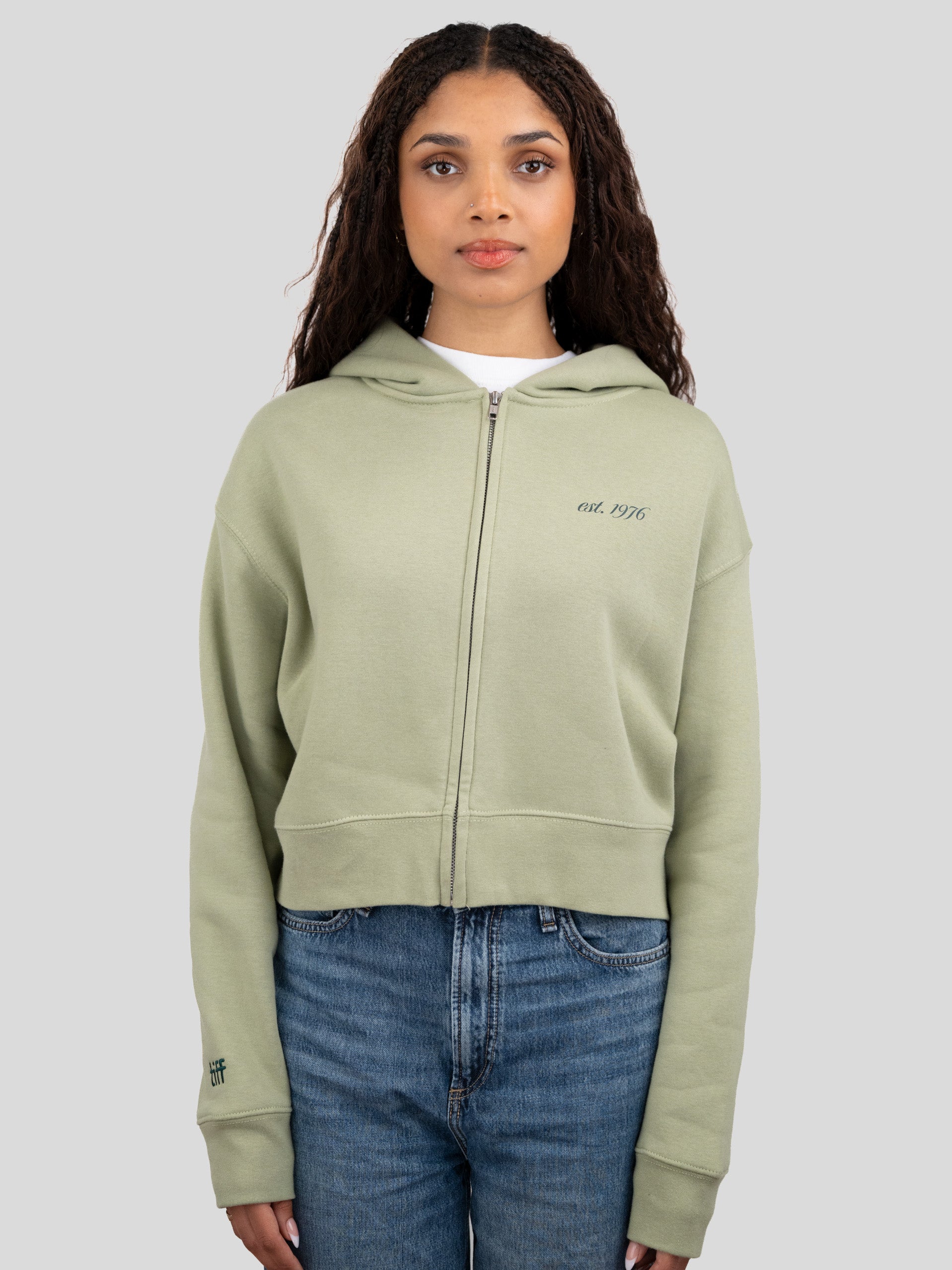 A woman modeling a light sage green cropped zip-up hoodie with 'est. 1976' embroidered on the left chest, paired with blue jeans, against a solid gray background.