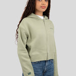 A woman wearing a light sage green cropped zip-up hoodie with a small embroidered logo on the chest and sleeve, paired with blue jeans, against a solid gray studio background.
