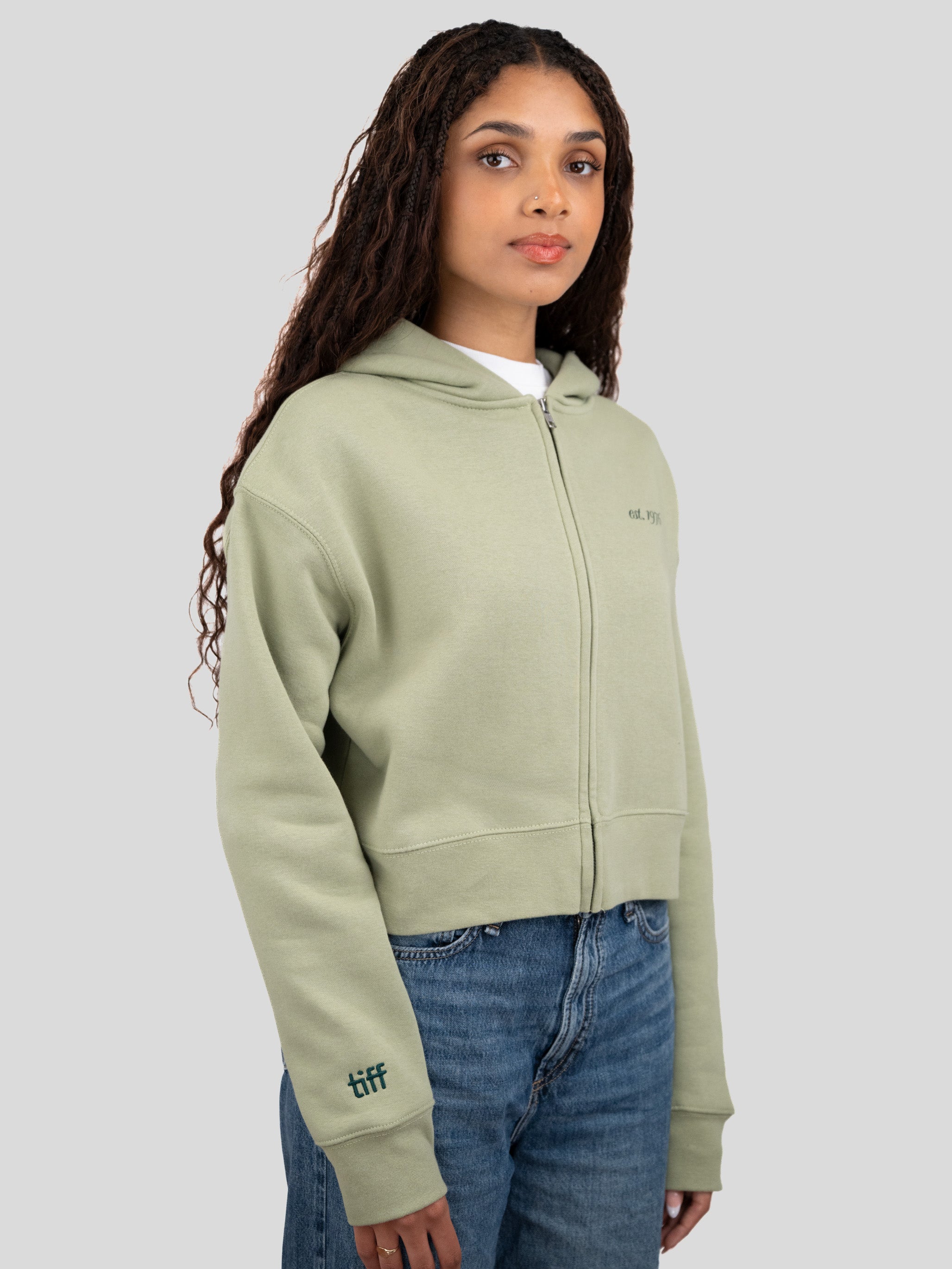 A woman wearing a light sage green cropped zip-up hoodie with a small embroidered logo on the chest and sleeve, paired with blue jeans, against a solid gray studio background.