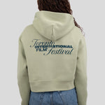 A model viewed from behind wearing a light sage green cropped hoodie with a dark green graphic print reading 'Toronto International Film Festival' on the back, paired with blue jeans against a solid gray background.