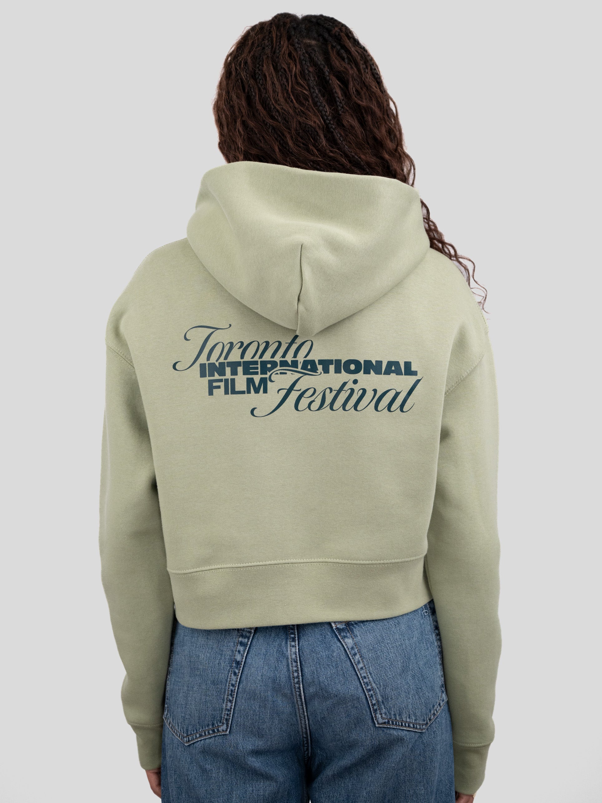 A model viewed from behind wearing a light sage green cropped hoodie with a dark green graphic print reading 'Toronto International Film Festival' on the back, paired with blue jeans against a solid gray background.