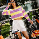A woman wearing a yellow and purple striped cropped long-sleeve shirt with a 'tiff' logo on the chest, white shorts, and white sunglasses, posing next to orange and black bicycles in an outdoor urban setting.