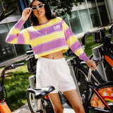 A woman wearing a yellow and purple striped cropped long-sleeve shirt with a 'tiff' logo on the chest, white shorts, and white sunglasses, posing next to orange and black bicycles in an outdoor urban setting.