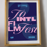 A framed art print featuring a stylized poster for the 2026 Toronto International Film Festival, displayed on a small round table against a light gray wall.
