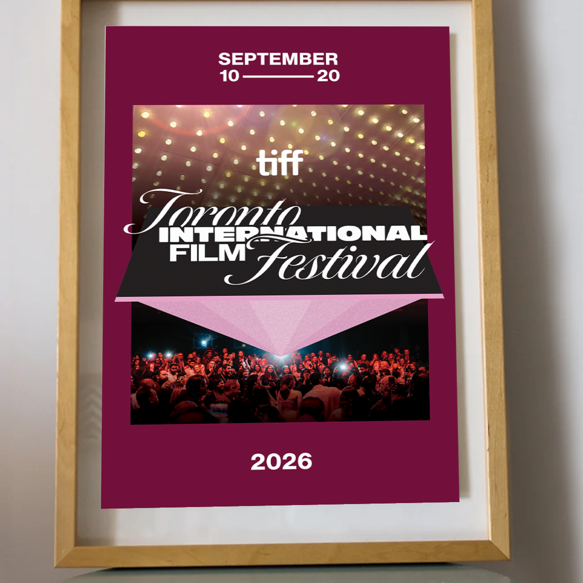 A framed poster for the 2026 Toronto International Film Festival (TIFF) leaning on a small round table against a light gray wall. The poster has a deep magenta background with white text, a photo of stage lights, and a photo of a film festival audience.
