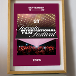 A framed poster for the 2026 Toronto International Film Festival (TIFF) leaning on a small round table against a light gray wall. The poster has a deep magenta background with white text, a photo of stage lights, and a photo of a film festival audience.
