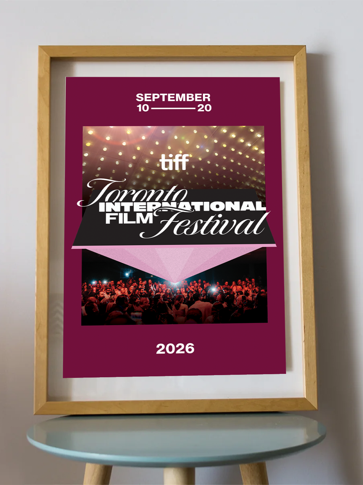 A framed poster for the 2026 Toronto International Film Festival (TIFF) leaning on a small round table against a light gray wall. The poster has a deep magenta background with white text, a photo of stage lights, and a photo of a film festival audience.