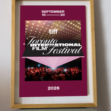 A framed poster for the 2026 Toronto International Film Festival (TIFF) leaning on a small round table against a light gray wall. The poster has a deep magenta background with white text, a photo of stage lights, and a photo of a film festival audience.