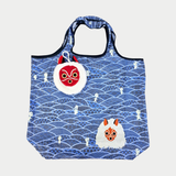 A blue reusable tote bag featuring a traditional geometric wave pattern dotted with small white Kodama spirits, an illustration of San's mask, and a matching plush mask pouch clipped to the handle.