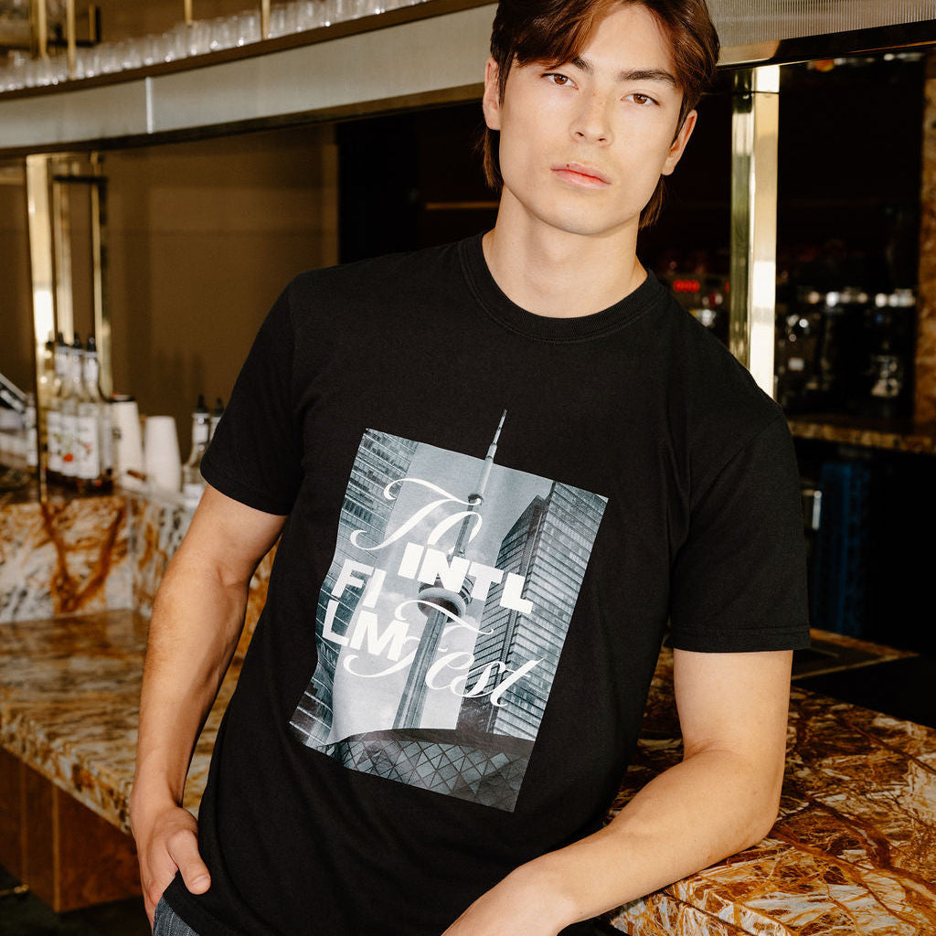 A man wearing a black graphic t-shirt with a New York City skyline print and gray striped pants, standing in a bar environment.