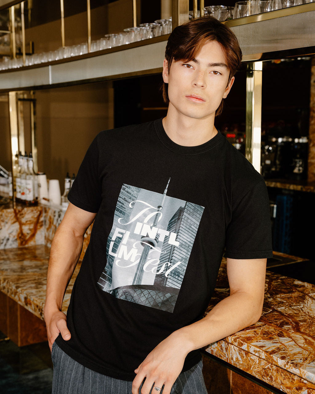 A man wearing a black graphic t-shirt with a New York City skyline print and gray striped pants, standing in a bar environment.