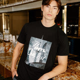 A man wearing a black graphic t-shirt with a New York City skyline print and gray striped pants, standing in a bar environment.